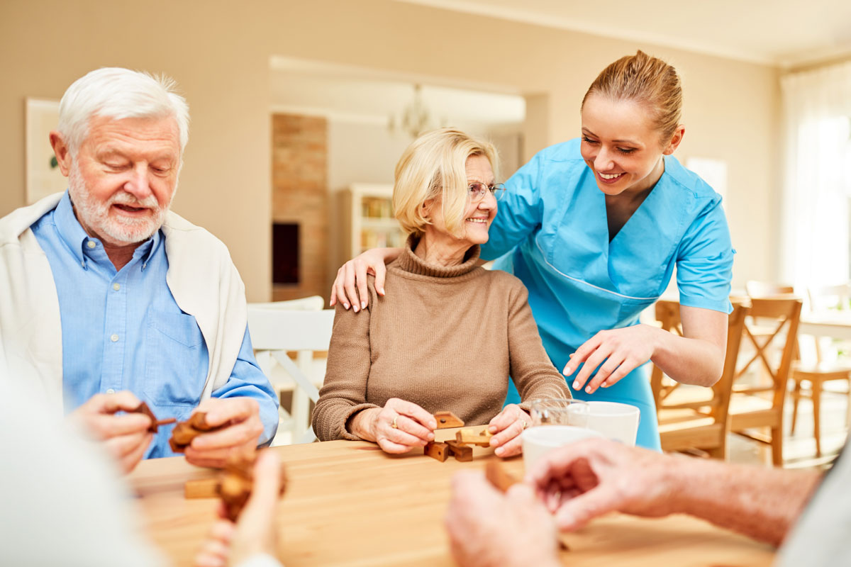 A staff member places her arm around a senior woman and talks with her as the woman sits at a table with other seniors working on wood puzzles