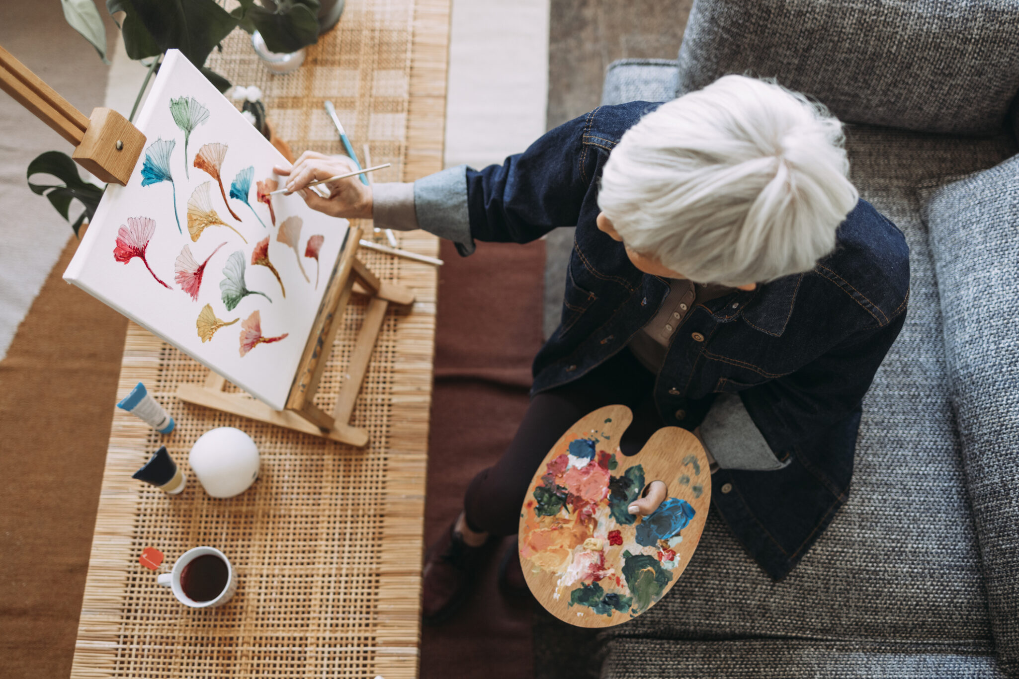 A top down photo of an older woman sitting on a couch and painting a flowers